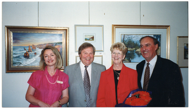 COL photo of four people, standing in a row, posing for a photo. They are all smiling. One of the women is holding a large bouquet of flowers. On the wall behind them, several paintings are displayed.