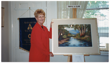 COL photo of a woman standing beside a painting on an easel. At the top of the easel is a small sign, with the text "TRIPLE A CLUB". Further down (below the painting) is another sign, with the text "RAFFLE PRIZE" (underlined), "In the Yarra Valley", "Donated by Barbara Ashford Wood", "$2 ticket". Hanging on a door behind the woman is a blue and gold 'Arthritis Victoria' banner. Beside the door (and behind the easel), there is a large curtained window.