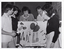 B&W photo of a small group of people standing around and behind a peg board, on which various items have been hung. One woman is pointing to an item on the board. At the bottom of the board is a sign that says, "ARTHRITIC AIDS".