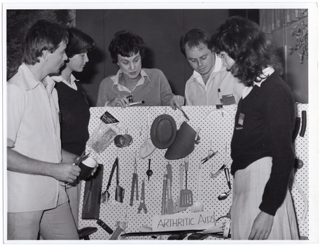 B&W photo of a small group of people standing around and behind a peg board, on which various items have been hung. One woman is pointing to an item on the board. At the bottom of the board is a sign that says, "ARTHRITIC AIDS".