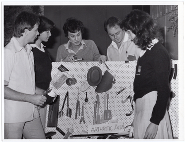 B&W photo of a small group of people standing around and behind a peg board, on which various items have been hung. One woman is pointing to an item on the board. At the bottom of the board is a sign that says, "ARTHRITIC AIDS".
