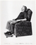 B&W photo of a man sitting in an lounge chair, smiling. The armchair has a padded headrest and armrests. On the floor in front of the armchair is a matching low footstool with four castor wheels. Written at the bottom of the photo is the text, "ARTISTIC AD REVERSE DIRECTION LEFT TO RIGHT”.