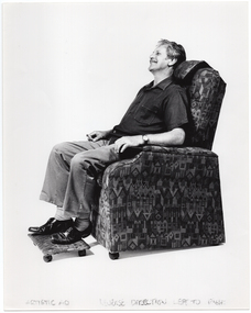 B&W photo of a man sitting in an lounge chair, smiling. The armchair has a padded headrest and armrests. On the floor in front of the armchair is a matching low footstool with four castor wheels. Written at the bottom of the photo is the text, "ARTISTIC AD REVERSE DIRECTION LEFT TO RIGHT”.