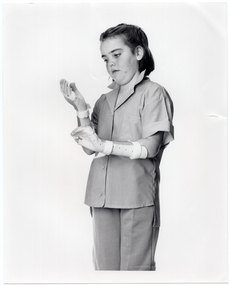 B&W photo of a girl wearing a hand and arm orthopaedic splint on each arm. She is looking down at her right hand and arm. She is wearing a short-sleeved shirt and trousers. Behind her is a plain background.