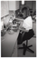 B&W photo of a woman sitting on a swivel chair on castors, at a kitchen sink. She is pouring water from a kettle into the sink. The kettle is sitting on a device which enables her to pour from the kettle without having to lift it. The sink is recessed into an L-shaped kitchen bench, upon which a cup and saucer can be seen sitting near the edge of the sink. There are several miscellaneous kitchen appliances on the bench. There are cupboards and drawers beneath the bench, and one set of cupboards above the sink.