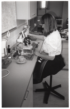 B&W photo of a woman sitting on a swivel chair on castors, at a kitchen sink. She is pouring water from a kettle into the sink. The kettle is sitting on a device which enables her to pour from the kettle without having to lift it. The sink is recessed into an L-shaped kitchen bench, upon which a cup and saucer can be seen sitting near the edge of the sink. There are several miscellaneous kitchen appliances on the bench. There are cupboards and drawers beneath the bench, and one set of cupboards above the sink.