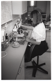 B&W photo of a woman sitting on a swivel chair on castors, at a kitchen sink. She is pouring water from a kettle into the sink. The kettle is sitting on a device which enables her to pour from the kettle without having to lift it. The sink is recessed into an L-shaped kitchen bench, upon which a cup and saucer can be seen sitting near the edge of the sink. There are several miscellaneous kitchen appliances on the bench. There are cupboards and drawers beneath the bench, and one set of cupboards above the sink.
