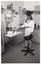 B&W photo of a woman standing beside a pull-out kitchen shelf and a high stool on castors. On top of the shelf are some kitchen appliances, such as a food processor and a hand-held immersion blender. On top of the kitchen cupboards, there are several round plastic food storage containers of various sizes stacked upon one another.
