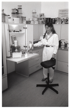 B&W photo of a woman standing beside a pull-out kitchen shelf and a high stool on castors. On top of the shelf are some kitchen appliances, such as a food processor and a hand-held immersion blender. On top of the kitchen cupboards, there are several round plastic food storage containers of various sizes stacked upon one another.
