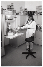 B&W photo of a woman standing beside a pull-out kitchen shelf and a high stool on castors. On top of the shelf are some kitchen appliances, such as a food processor and a hand-held immersion blender. On top of the kitchen cupboards, there are several round plastic food storage containers of various sizes stacked upon one another.