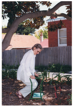 COL photo of a woman, from an upright position, pushing her arms down onto the armrests of a garden kneeler mobility aid. Her knees are bent at a 90 degree angle. She is either lowering herself down to a kneeling position or getting back up from a kneeling to a standing position.