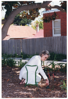 COL photo of a woman sitting back on her calves and feet while kneeling on a 'garden kneeler' mobility aid. She is holding a small garden fork or trowel in one hand, which she has dug into the ground. There are two other garden forks or trowels pushed into the ground in front of her. She is positioned beneath a large tree. In the background, some houses are visible from behind a wooden fence.