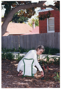 COL photo of a woman sitting back on her calves and feet while kneeling on a 'garden kneeler' mobility aid. She is holding a small garden fork or trowel in one hand, which she has dug into the ground. There are two other garden forks or trowels pushed into the ground in front of her. She is positioned beneath a large tree. In the background, some houses are visible from behind a wooden fence.