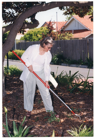 COL photo of a woman standing upright and leaning forward slightly. She is holding a long-handled gardening two-pronged tool which she uses to till the soil.