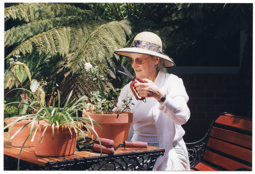 COL photo of a woman is sitting at a table in the garden. She is holding up a pair of gardening shears with thick handles. On the table, there are several potted plants and three more pairs of various types of small gardening tools, all with the same type of thick handles.