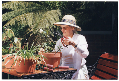 COL photo of a woman is sitting at a table in the garden. She is holding up a pair of gardening shears with thick handles. On the table, there are several potted plants and three more pairs of various types of small gardening tools, all with the same type of thick handles.