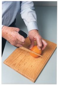 COL close up photo of a man's hands using a knife with a specially-designed handle. He is cutting an orange on a wooden chopping board.