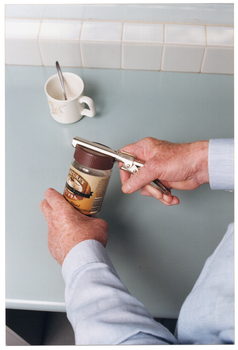 COL close up photo of a man's hands using a jar-opener to open the plastic lid of a glass coffee jar. He is standing at a kitchen bench. On the benchtop, there is a mug with a spoon in it.