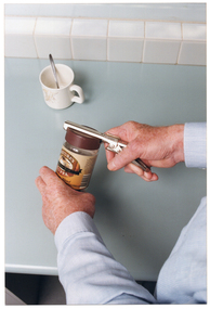 COL close up photo of a man's hands using a jar-opener to open the plastic lid of a glass coffee jar. He is standing at a kitchen bench. On the benchtop, there is a mug with a spoon in it.