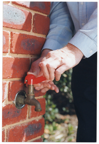 COL close up photo of a man using a tap-turner tool to open a garden tap. The tap is located on an exterior brick wall.