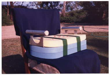 COL close up photo of a chair (without armrests) which has been adapted so that the seat is raised to a higher position. Three swimming pool kickboards (made of dense foam) have been stacked on top of one another, and a kitchen tray with handles has been placed between the top and second kickboards. The stack of kickboards and tray have been taped together and the entire thing has been placed on the seat of the chair. The chair has been placed outdoors. In the background, a gravel area and some trees are visible.