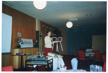 COL photo of a woman holding up a shower stool mobility aid. Behind her is a toilet chair mobility aid. Beside her is a table and a slide projecter. Behind her is another table with some gym hand weights, a large Arthritis Victoria sign, and a projector screen on the wall. Tables and chairs are visible in the room, which is a large hall.