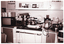 B&W photo of some saucepans, a kettle, a plate and carry tray, canisters, and other items commonly found in a kitchen are placed next to or stacked on top of one another on a kitchen bench and stove. Also on the kitchen bench are a small portable television set and a small benchtop oven. Above the bench and stove is a small shelf beneath some wall-mounted cupboards. On the shelf are two small clocks, a photo frame or mirror, and some ornaments.