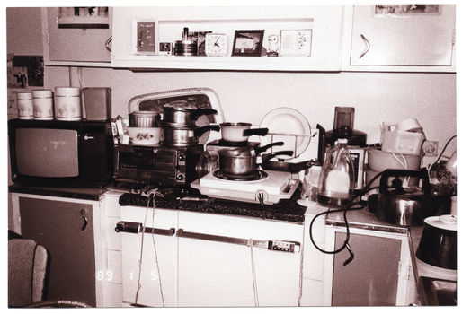 B&W photo of some saucepans, a kettle, a plate and carry tray, canisters, and other items commonly found in a kitchen are placed next to or stacked on top of one another on a kitchen bench and stove. Also on the kitchen bench are a small portable television set and a small benchtop oven. Above the bench and stove is a small shelf beneath some wall-mounted cupboards. On the shelf are two small clocks, a photo frame or mirror, and some ornaments.