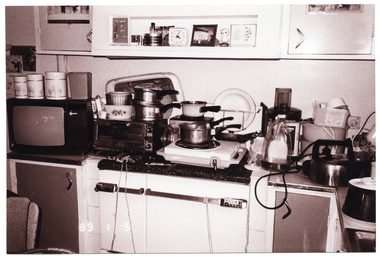 B&W photo of some saucepans, a kettle, a plate and carry tray, canisters, and other items commonly found in a kitchen are placed next to or stacked on top of one another on a kitchen bench and stove. Also on the kitchen bench are a small portable television set and a small benchtop oven. Above the bench and stove is a small shelf beneath some wall-mounted cupboards. On the shelf are two small clocks, a photo frame or mirror, and some ornaments.