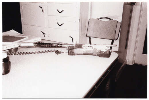 B&W photo of a telephone receiver, strapped to a long rod with tape, on a table amongst other items. At one end of the table is an empty chair, with a cupboard with drawers behind it. The photo is date-stamped "89 1 5" in white text on the bottom left side of the image layer (although, on this photo, the date stamp is barely noticeable and difficult to decipher against the pale coloured table).