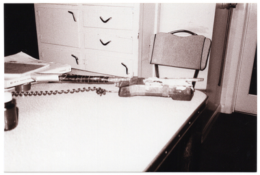 B&W photo of a telephone receiver, strapped to a long rod with tape, on a table amongst other items. At one end of the table is an empty chair, with a cupboard with drawers behind it. The photo is date-stamped "89 1 5" in white text on the bottom left side of the image layer (although, on this photo, the date stamp is barely noticeable and difficult to decipher against the pale coloured table).