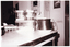 B&W photo of an insulated teapot and some kitchen canisters amongst various other items on a kitchen table. The photo is date-stamped "89 1 5" in white text on the bottom left side of the image layer.