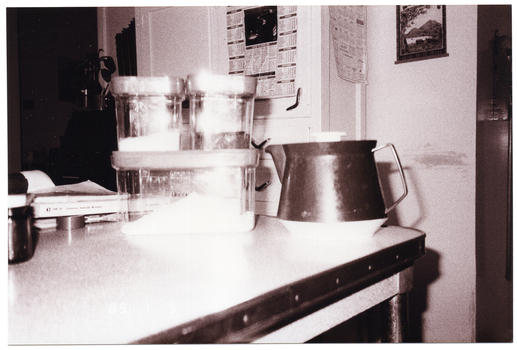 B&W photo of an insulated teapot and some kitchen canisters amongst various other items on a kitchen table. The photo is date-stamped "89 1 5" in white text on the bottom left side of the image layer.