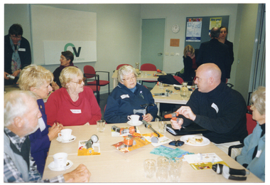 COL photo of a small group of people sitting around a table, on which there are various garden hose nozzle attachments. A man is demonstrating the use of one of the hose attachments. Behind them, there are other people are either sitting at tables or standing.