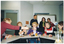 COL photo of a small group of people sitting around some tables set up in a circle. The tables have various garden hose nozzle attachments on them, which some people are passing to one another. Behind them, three people are standing in front of a whiteboard. One of them is holding up a thick stack of colour-printed leaflets.