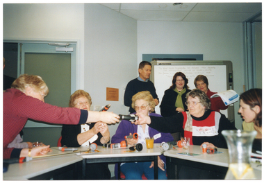COL photo of a small group of people sitting around some tables set up in a circle. The tables have various garden hose nozzle attachments on them, which some people are passing to one another. Behind them, three people are standing in front of a whiteboard. One of them is holding up a thick stack of colour-printed leaflets.