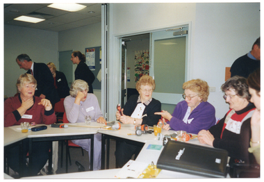 COL photo of a small group of people sitting around some tables set up in a circle. The tables have various garden hose nozzle attachments on them, which some people are looking at or holding. In the background, on either side of an open doorway, a few other people are standing around in the room behind them.