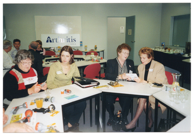 COL photo of a small group of people sitting around some tables set up in a circle. The tables have various garden hose nozzle attachments on them. Some people are looking at the garden hose nozzles while others are looking at some brochures. There are also some glasses of orange juice and water and a carafe of water on the table. Behind the group, a few other people are seated at another group of tables. There is a large whiteboard on the wall with an Arthritis Victoria sign sitting on the bottom ledge, leaning back against the whiteboard.