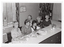 B&W photo of a small group of women sitting on one side of a long table. On the table, there are some plastic containers with lids and a small tray of sandwiches, some jugs of milk and sugar bowls, a couple of cups, and two books. On the wall behind them is a small sign with the handwritten words, 'The Rheumatism and Arthritis Association of Victoria'. To one side of the sign, there is a stained glass window and, below that, a radiator with a tea towel draped across the top.