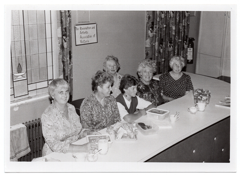 B&W photo of a small group of women sitting on one side of a long table. On the table, there are some plastic containers with lids and a small tray of sandwiches, some jugs of milk and sugar bowls, a couple of cups, and two books. On the wall behind them is a small sign with the handwritten words, 'The Rheumatism and Arthritis Association of Victoria'. To one side of the sign, there is a stained glass window and, below that, a radiator with a tea towel draped across the top.
