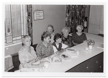 B&W photo of a small group of women sitting on one side of a long table. On the table, there are some plastic containers with lids and a small tray of sandwiches, some jugs of milk and sugar bowls, a couple of cups, and two books. On the wall behind them is a small sign with the handwritten words, 'The Rheumatism and Arthritis Association of Victoria'. To one side of the sign, there is a stained glass window and, below that, a radiator with a tea towel draped across the top.