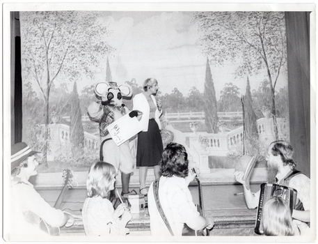 B&W photo of a woman and a person dressed in a rat costume, standing on a stage. Behind them is a painted backdrop depicting a large garden enclosed by white Roman balustrades. There is an accordion and an accordion case on the stage floor. In the foreground, a group of four musicians are standing on the floor below the stage. They are holding their instruments. One of the musicians is holding his hat in one hand.