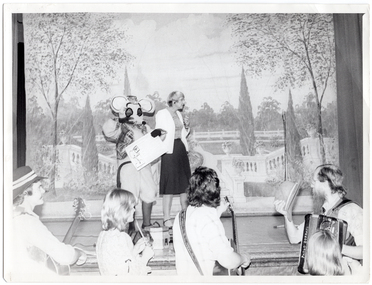 B&W photo of a woman and a person dressed in a rat costume, standing on a stage. Behind them is a painted backdrop depicting a large garden enclosed by white Roman balustrades. There is an accordion and an accordion case on the stage floor. In the foreground, a group of four musicians are standing on the floor below the stage. They are holding their instruments. One of the musicians is holding his hat in one hand.