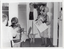 B&W photo of a woman and a mascot dressed in a rat costume standing in the corner of a room. There is a gas wall heater on the wall beside them, and a small window behind them. The mascot is holding a large piece of cardboard: on one half, there is a poster depicting a beach scene with a tall palm tree, and the text, "Beautiful MALAYSIA"; the other half contains the text, "BUY A TICKET FOR RAAV". There is a small table and chair in front of the wall heater. In the foreground, there are several people seated on chairs.