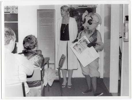 B&W photo of a woman and a mascot dressed in a rat costume standing in the corner of a room. There is a gas wall heater on the wall beside them, and a small window behind them. The mascot is holding a large piece of cardboard: on one half, there is a poster depicting a beach scene with a tall palm tree, and the text, "Beautiful MALAYSIA"; the other half contains the text, "BUY A TICKET FOR RAAV". There is a small table and chair in front of the wall heater. In the foreground, there are several people seated on chairs.