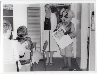 B&W photo of a woman and a mascot dressed in a rat costume standing in the corner of a room. There is a gas wall heater on the wall beside them, and a small window behind them. The mascot is holding a large piece of cardboard: on one half, there is a poster depicting a beach scene with a tall palm tree, and the text, "Beautiful MALAYSIA"; the other half contains the text, "BUY A TICKET FOR RAAV". There is a small table and chair in front of the wall heater. In the foreground, there are several people seated on chairs.