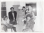 B&W photo of a small group of people, dressed in evening wear. They are gathered around a small table. Among them is a person dressed in a rat costume. Most of the people are standing behind a small table, while a few others are seated around the table. One woman, who is sitting, has one leg up on the table. Everyone is smiling or laughing. There is a gas wall heater on the wall behind them, with two small windows on either side.