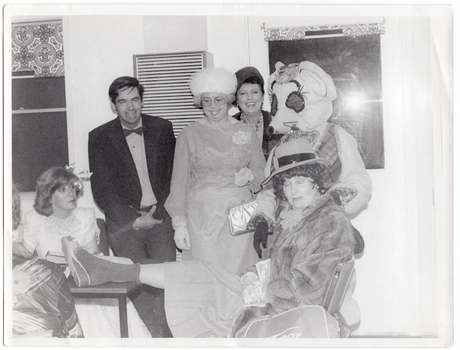B&W photo of a small group of people, dressed in evening wear. They are gathered around a small table. Among them is a person dressed in a rat costume. Most of the people are standing behind a small table, while a few others are seated around the table. One woman, who is sitting, has one leg up on the table. Everyone is smiling or laughing. There is a gas wall heater on the wall behind them, with two small windows on either side.