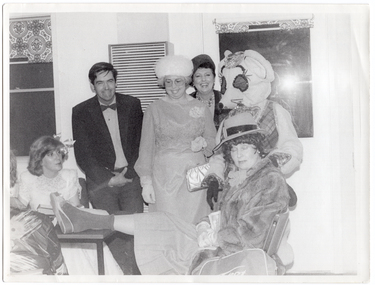 B&W photo of a small group of people, dressed in evening wear. They are gathered around a small table. Among them is a person dressed in a rat costume. Most of the people are standing behind a small table, while a few others are seated around the table. One woman, who is sitting, has one leg up on the table. Everyone is smiling or laughing. There is a gas wall heater on the wall behind them, with two small windows on either side.