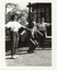 B&W photo of three people conducting in an exercise demonstration on the grounds of a building (which is behind them). In the middle is a man wearing a suit, shirt and tie. On either side of him are two women wearing t-shirts printed with the logo, "Joint Action", and track suit pants.